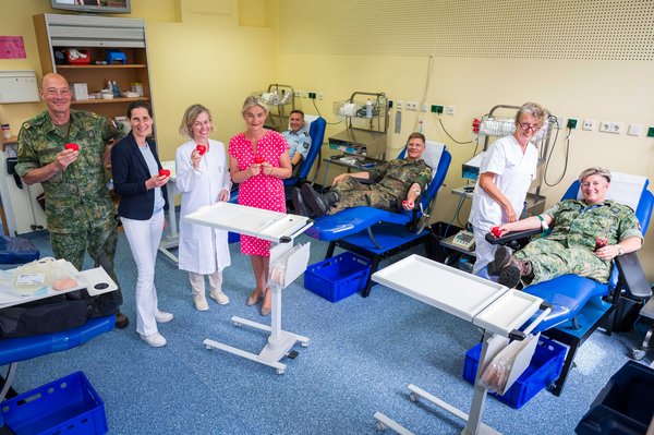 UKM | Ein Mann in Polizeiuniform und zwei Menschen im Feldanzug auf Liegen in der Blutspendeabteilung. Dazwischen steht eine Pflegerin. Außerdem stehen im Raum: ein Mann im Feldanzug und drei Frauen halten kleine rote Plastikherzen in den Händen. Alle lächeln.